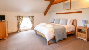 A bedroom with a wooden ceiling, a bed with white bedding, a nightstand with a lamp, and a window with floral curtains. - Woodthorpe Cruck Cottage (Chesterfield)