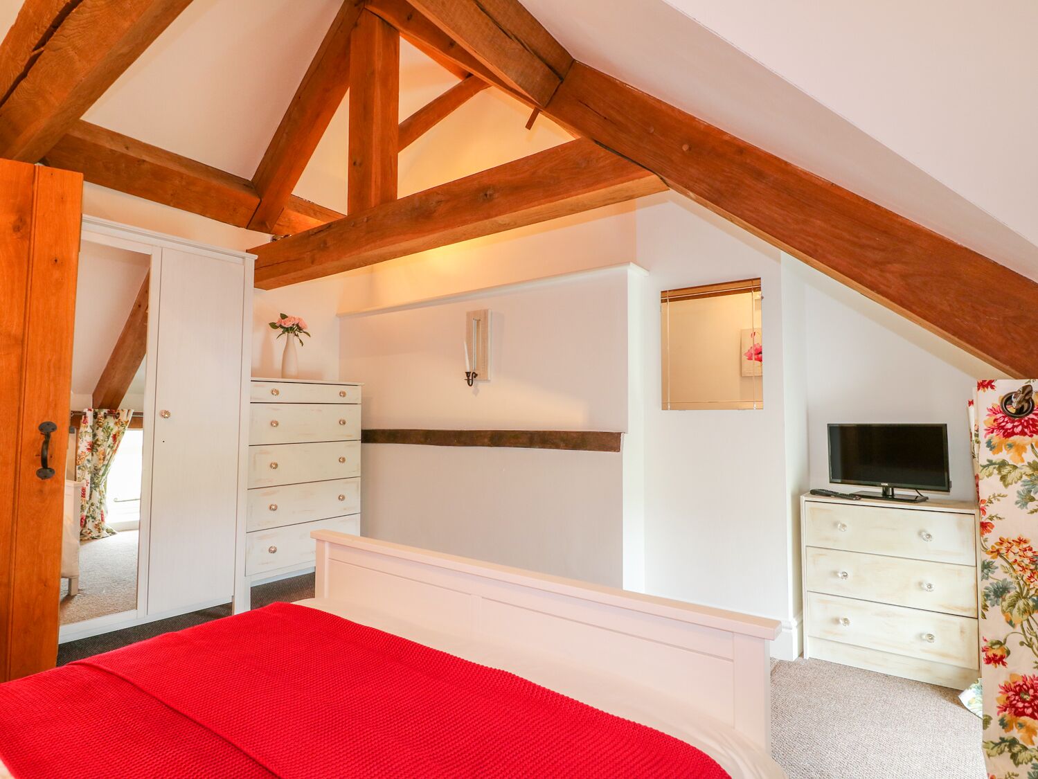Une chambre avec un plafond en pente, des poutres en bois, un lit recouvert d’une couverture rouge, une commode, un téléviseur et un miroir.