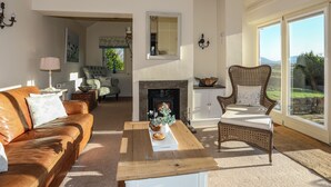 Un salon avec une cheminée, un canapé, un fauteuil en osier et une vue sur l’extérieur.