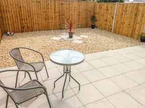 Un espace patio avec une table ronde et deux chaises en osier, entouré d’une clôture en bois et de galets.