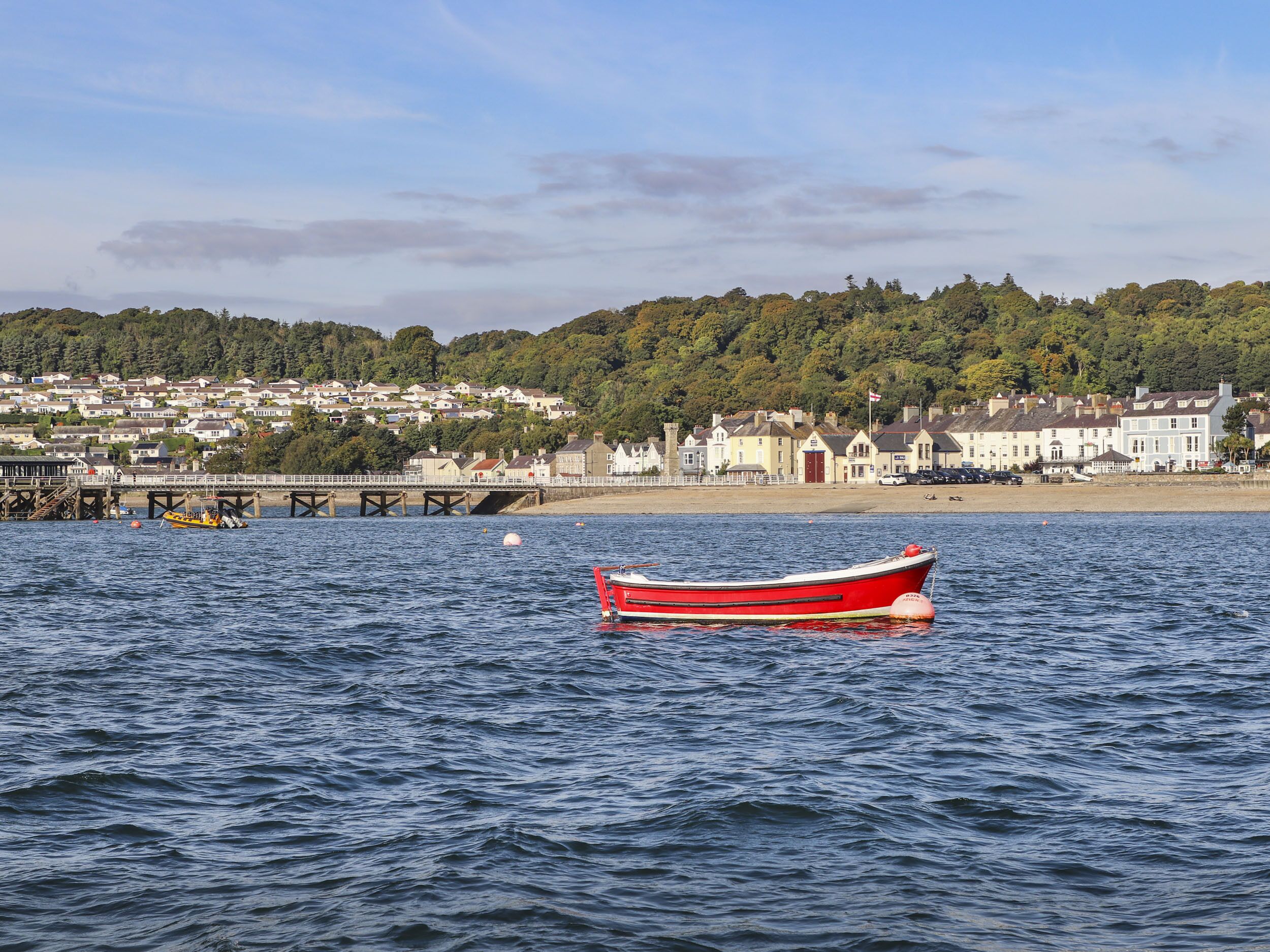 Un bateau rouge, amarré dans des eaux calmes, avec en arrière-plan une ville côtière et une colline boisée.