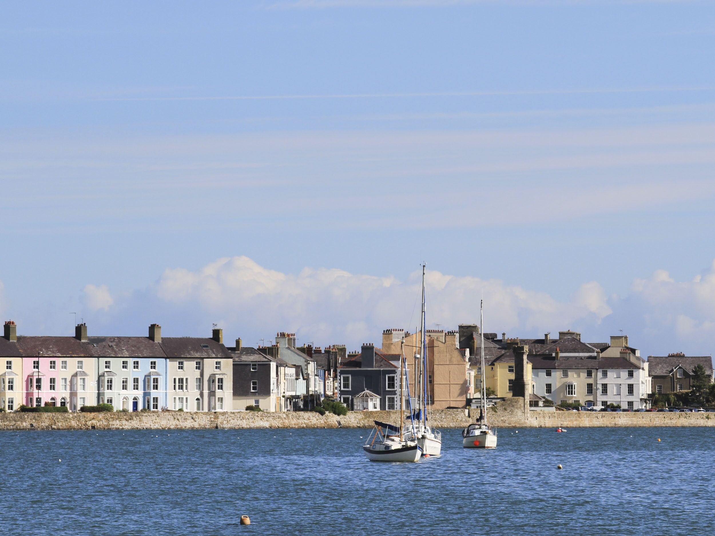Eine Küstenstadt mit bunten Häusern entlang der Uferlinie, zwei Segelboote im Wasser und einem klaren Himmel.