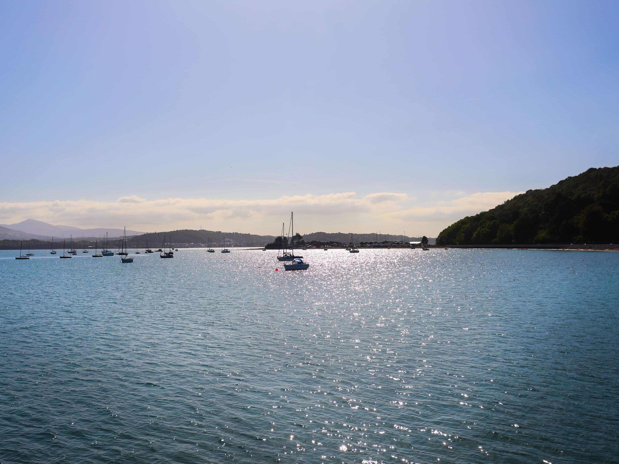 Ein ruhiger See mit mehreren Segelbooten, die vor Anker liegen, umgeben von Hügeln und blauem Himmel.
