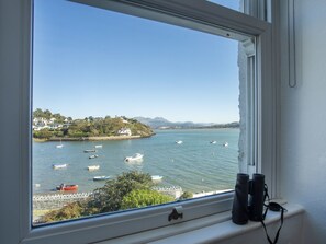Une vue sur la côte, aperçue à travers une fenêtre, avec des jumelles posées sur le rebord, donnant sur des eaux calmes où flottent plusieurs bateaux, et sur une côte lointaine parsemée de maisons.