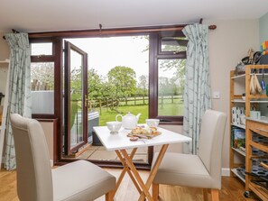 Un coin repas accueillant avec une table dressée pour deux personnes, un espace cuisine équipé d’ustensiles et une vue sur un paysage verdoyant grâce à de grandes fenêtres.