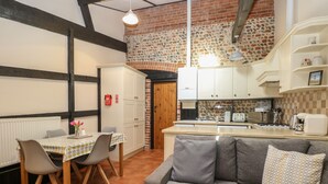 A cozy kitchen with a stone backsplash, a dining area with a table and chairs, and a grey sofa with patterned cushions. - Hitchens Cottage (Norwich)