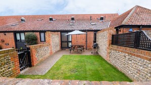 A brick house with a tiled roof, a small lawn, and a patio area with a table and chairs. - Hitchens Cottage (Norwich)
