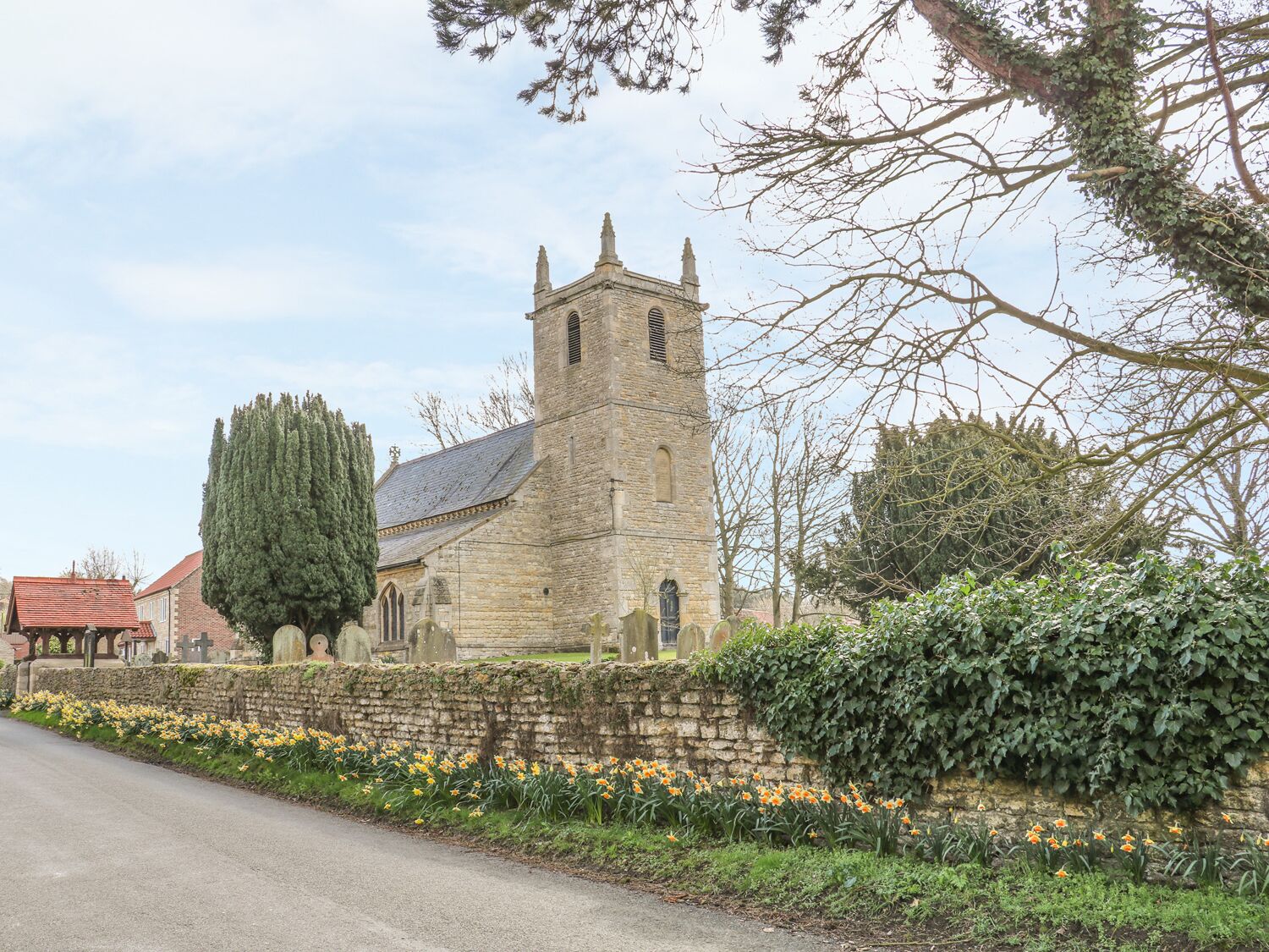 Una iglesia de piedra con un alto campanario, un árbol grande y una jardinera a lo largo de la carretera.