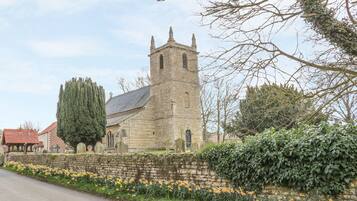 Una iglesia de piedra con un alto campanario, un árbol grande y una jardinera a lo largo de la carretera.