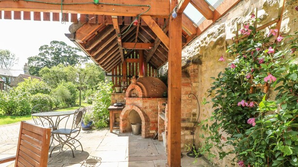 An outdoor patio with a brick oven, wooden furniture, and a garden wall. - Forge Cottage (Gainsborough)