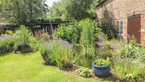 A garden with a pond, various plants, and a brick building with a window and door. - Forge Cottage (Gainsborough)
