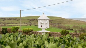 A white birdhouse with two birds perched on it, surrounded by green plants and a stone wall, set against a backdrop of rolling hills and a clear sky. - The Doves (Bridport)