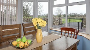 A conservatory with a table set for breakfast, featuring a fruit bowl, a vase with yellow flowers, and a view of a stone wall and green field. - Plas Cefn Mawr (Llanfairpwllgwyngyll)