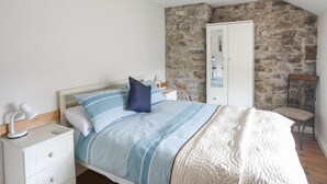 A bedroom with a bed, nightstand, lamp, and a stone wall. - Plas Cefn Mawr (Llanfairpwllgwyngyll)