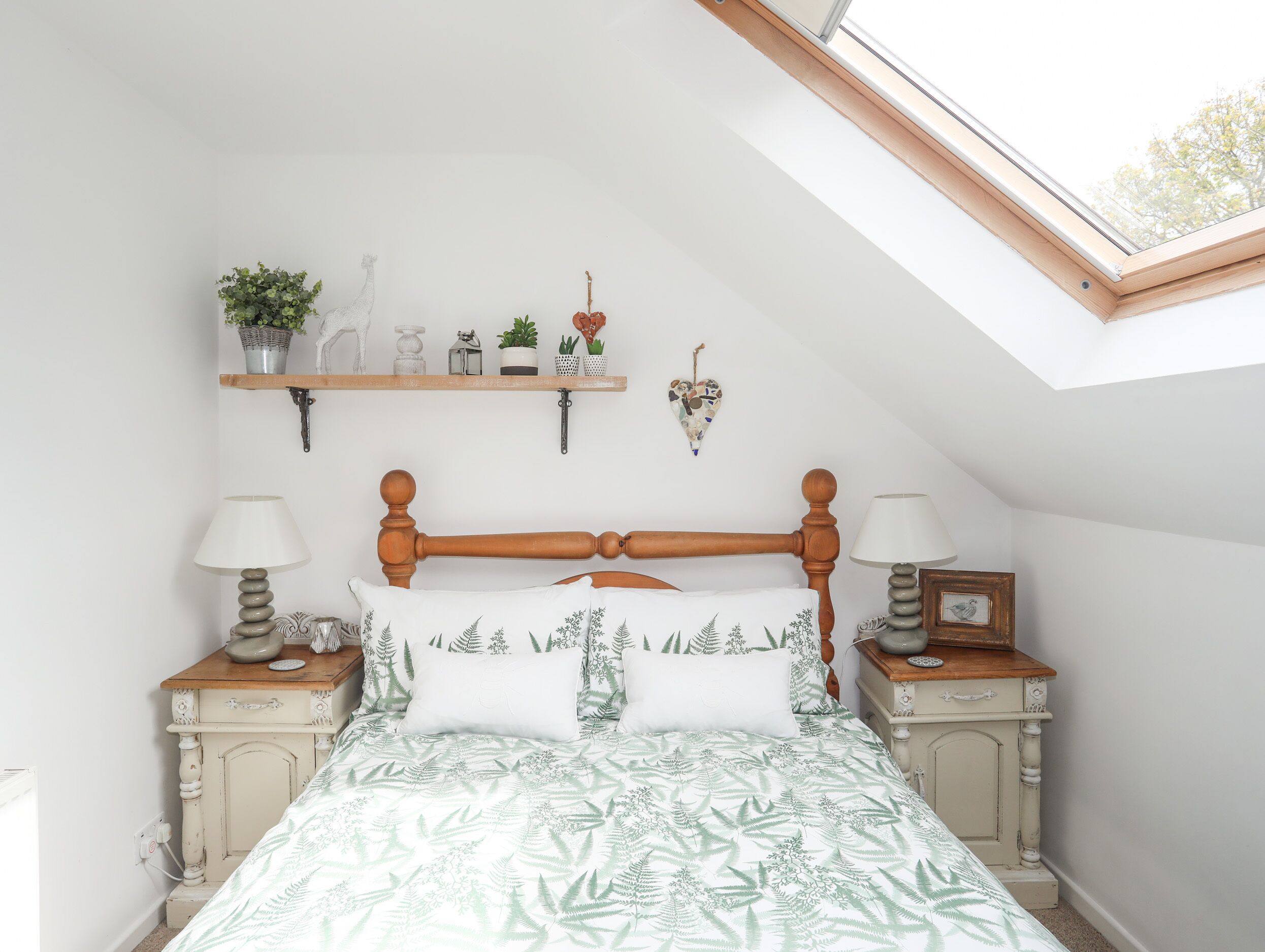 A bedroom with a sloped ceiling, a wooden bed frame, a green leaf-patterned bedspread, bedside tables with lamps, and a skylight.