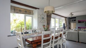 Un espace repas avec une table en bois et des chaises blanches, un lustre et une vue sur la verdure à l’extérieur.
