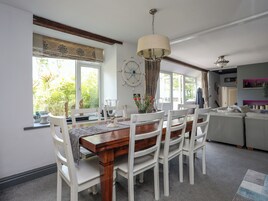 Uma sala de jantar com mesa de madeira e cadeiras brancas, um lustre e vista para o verde lá fora.