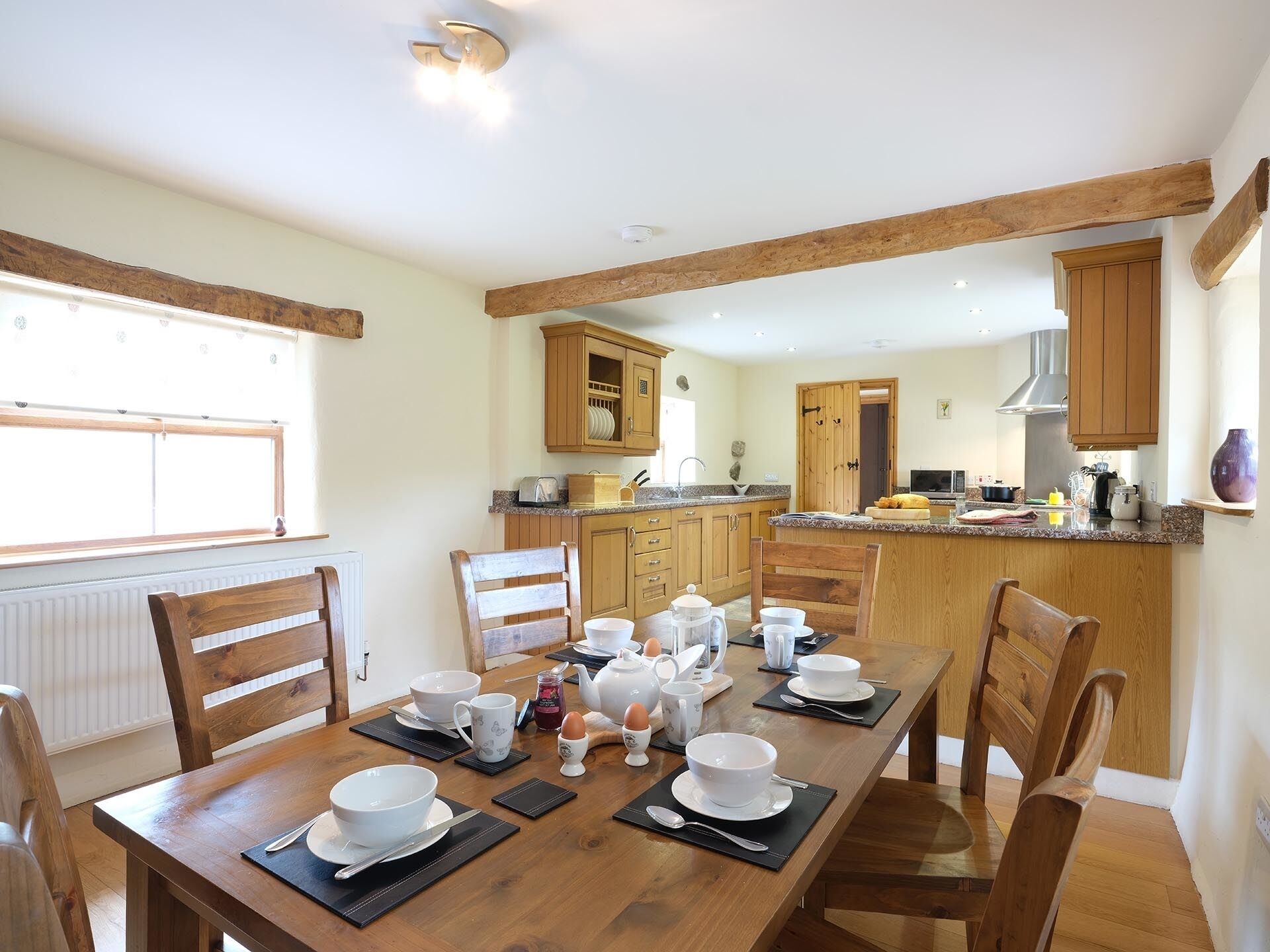 A dining area with a wooden table set for breakfast, wooden chairs, and a kitchen in the background.