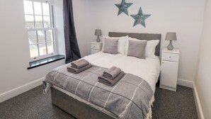 Quarto com cama, duas mesas de cabeceira, janela com cortinas e dois detalhes decorativos em formato de estrela na parede.