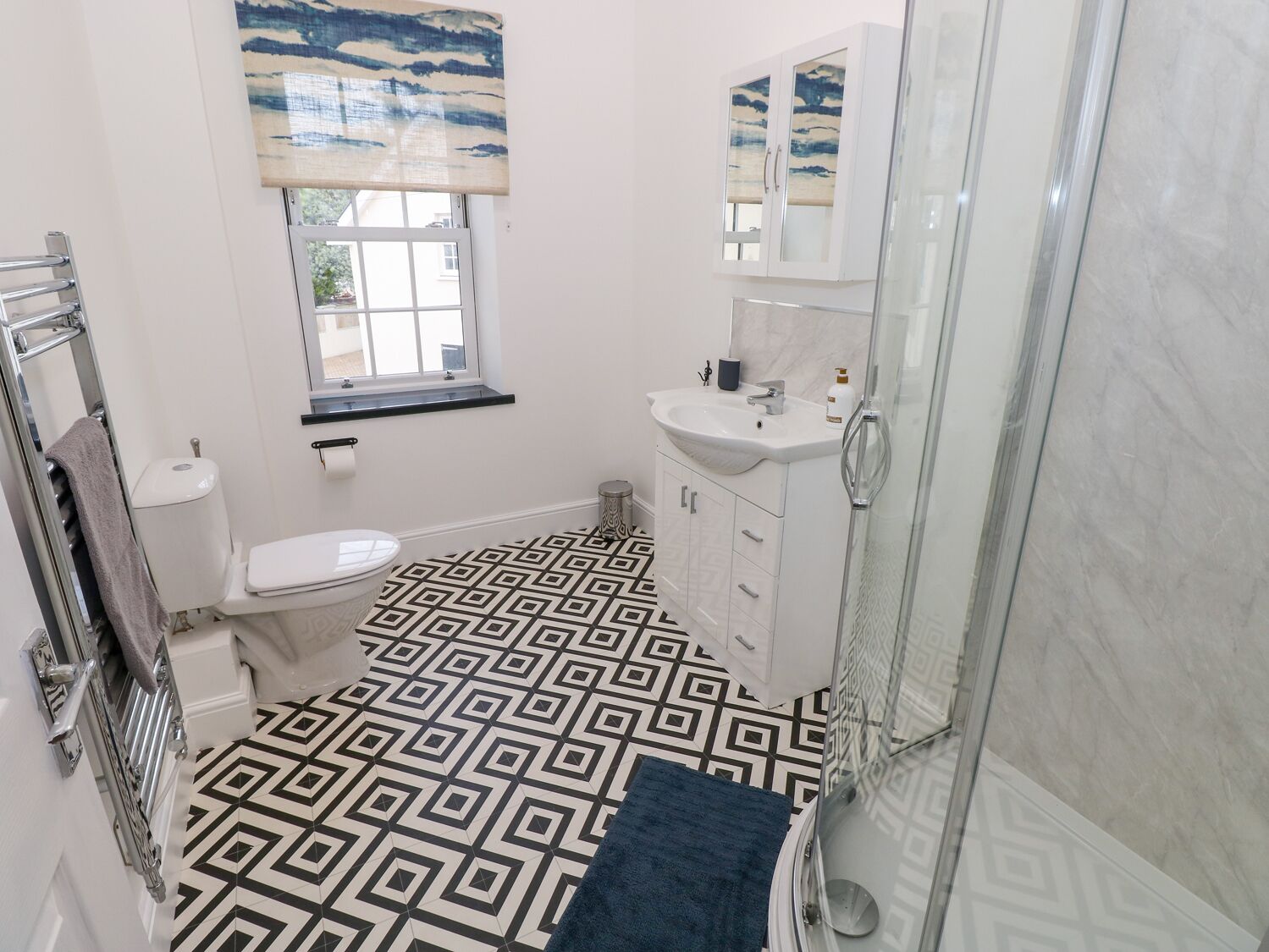 A bathroom with a geometric patterned floor, a toilet, a sink, a cabinet, a window with a view, and a shower area.