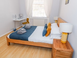 A neatly made bed with a blue blanket, pillows, and folded towels. There is a wooden nightstand with a lamp, a small table with a lamp, and a chair.