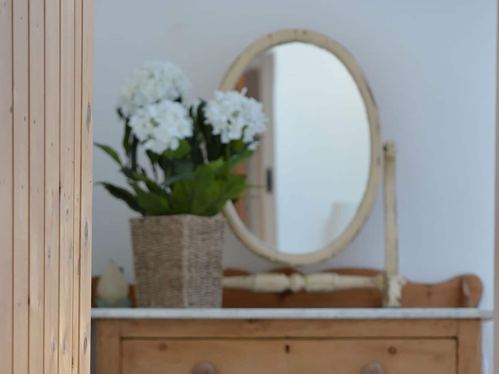 Une commode en bois avec un miroir et un panier en osier contenant des fleurs blanches.