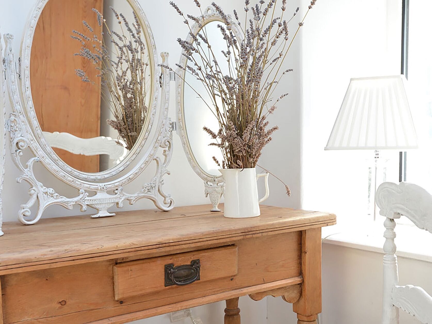 A wooden dresser with two ornate mirrors, a white vase with dried lavender, and a white lamp.