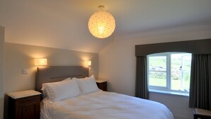 A bedroom with a large bed, bedside lamps, a nightstand, a window with curtains, and a view of the outdoors. - Culverwells (Axminster)