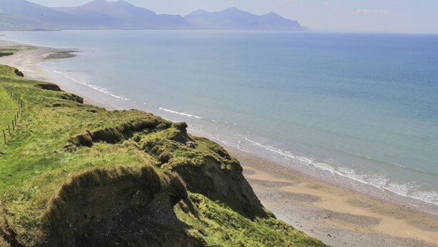 A coastal landscape with a grassy cliff, beach, and distant mountains under a partly cloudy sky.