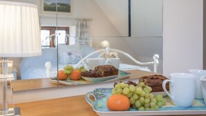 A breakfast tray with grapes, an orange, and cookies on a table with a lamp and a mirror reflecting a bed and a window.