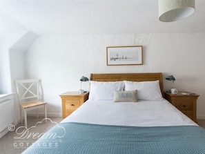 A bedroom with a wooden bed, white bedding, a framed picture on the wall, and a small table with a lamp.