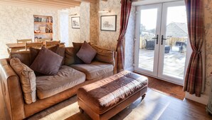 A cozy living room with a brown leather sofa, a matching ottoman, and a dining area with a wooden table and chairs. There is a bookshelf with books and framed pictures on the wall, and a sliding glass door leading to a patio with a wooden fence. - The Pheasantry (Scarborough)