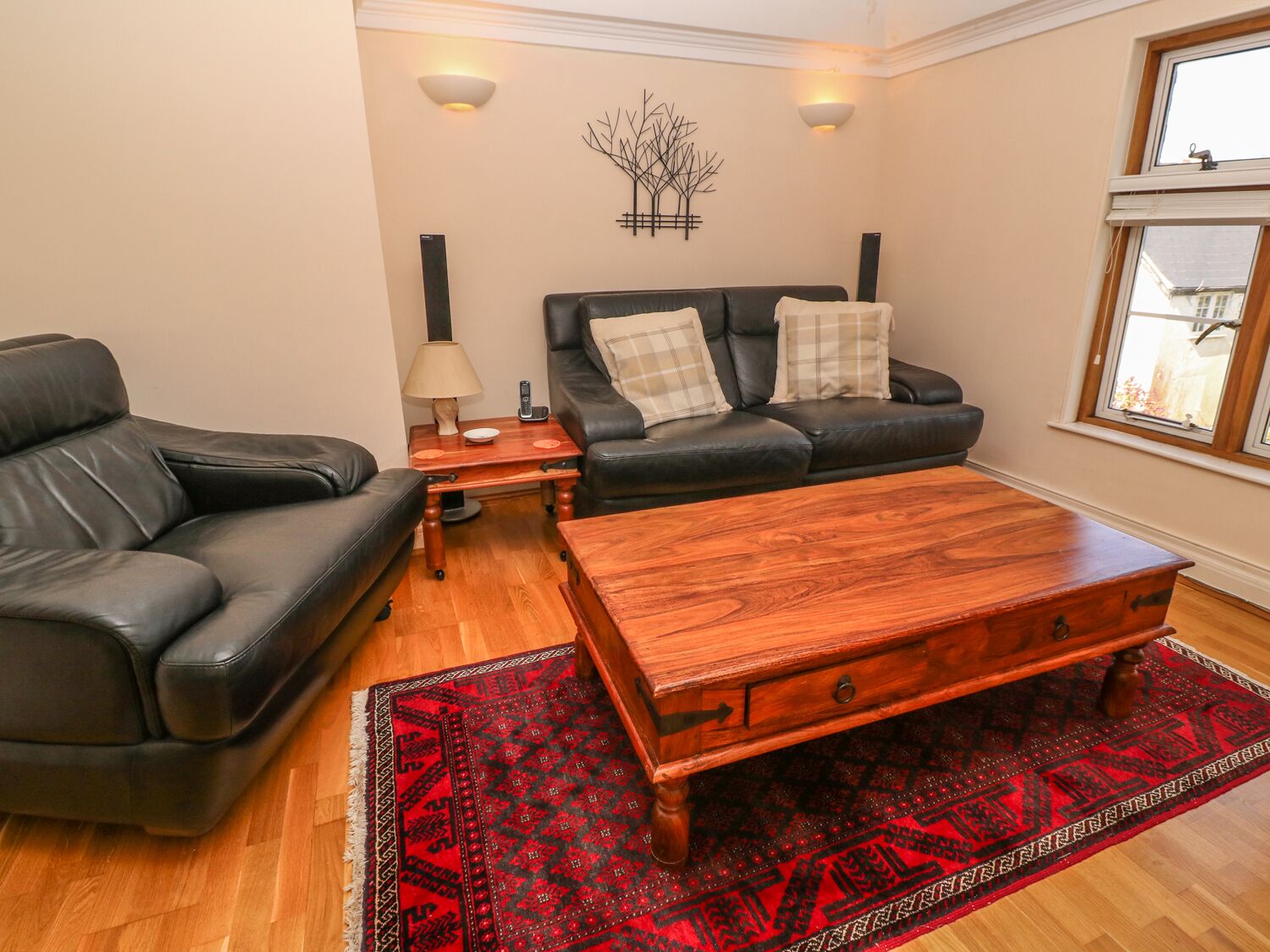Un salon avec un canapé en cuir noir, une table basse en bois, un tapis à motifs rouges et deux lampes fixées au mur.