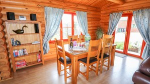A cozy cabin interior with a wooden dining table, chairs, a bookshelf, and a window with curtains. - Yew Lodge (Oakham)
