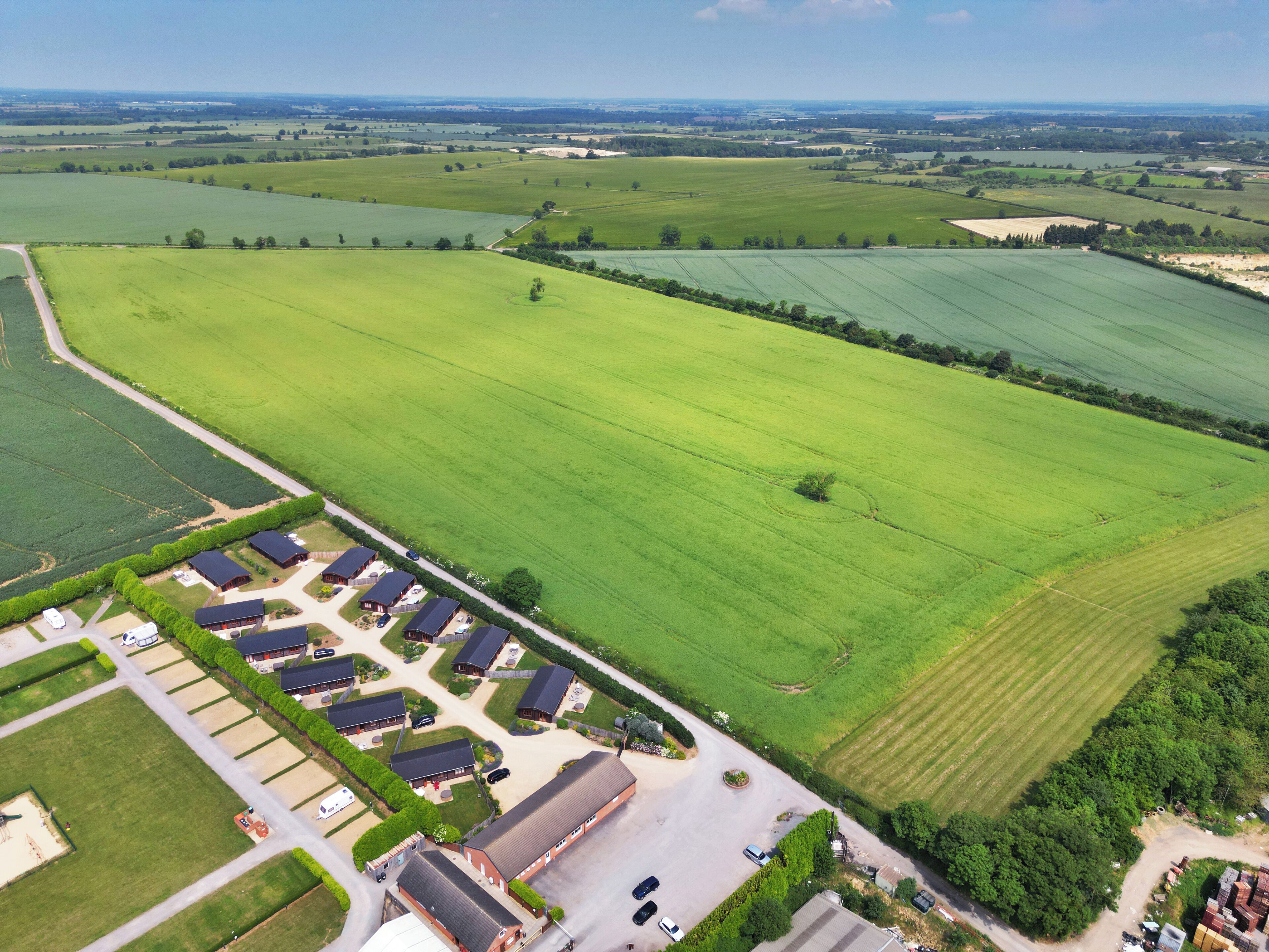 Vista aérea de uma área rural com um grande campo verde, uma fileira de casas com telhados escuros e um espaço para estacionamento.