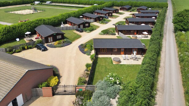 A rural setting with multiple wooden cabins arranged in a row along a road, surrounded by green fields and a clear sky. - Yew Lodge (Oakham)