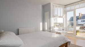 A bedroom with a large bed, a radiator, a chair, and a balcony with a view of buildings. - Chy-an-brae (Newquay)