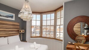 A bedroom with a large bed, a wooden headboard, a chandelier, a window with blinds, a round mirror, and a desk with a chair. - Harbourside House (Weymouth)
