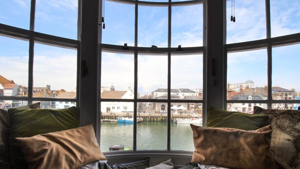 A bay window with a view of a harbor, a cushioned bench, and a patterned cushion. - Harbourside House (Weymouth)