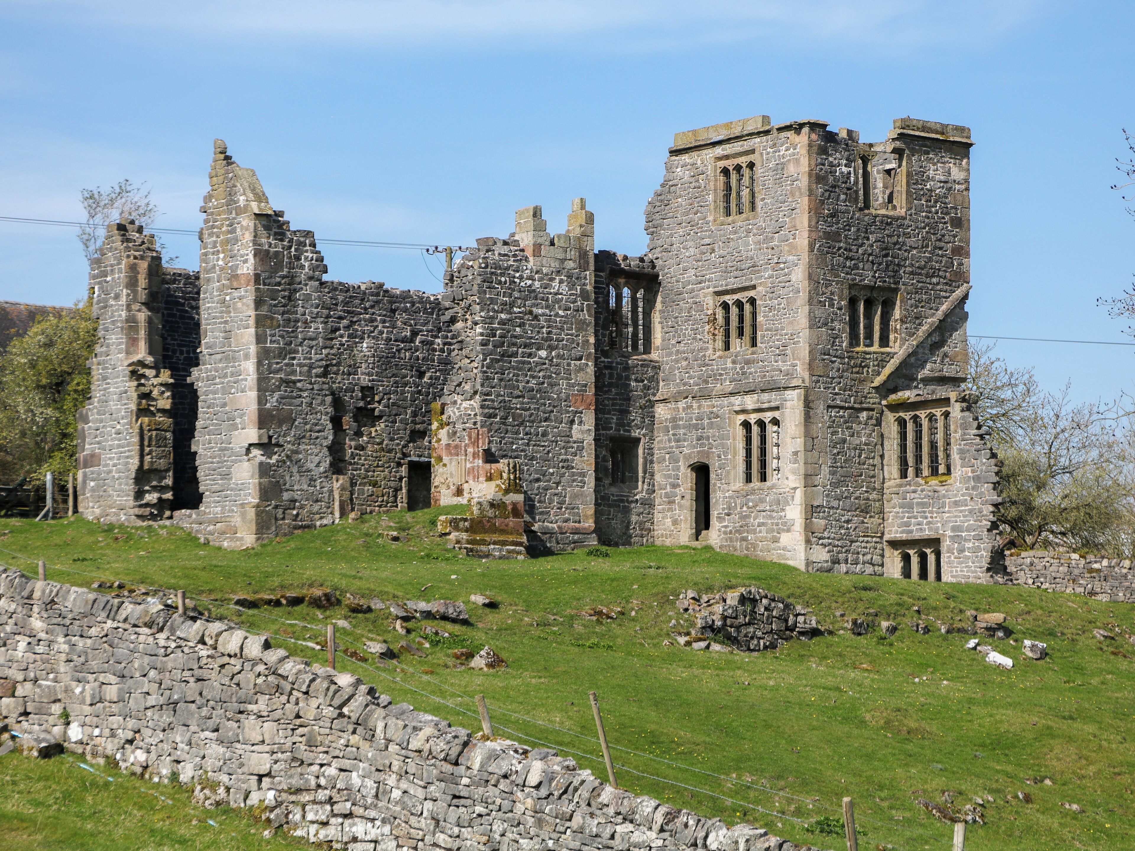 A historic stone building with multiple towers and arched windows, surrounded by a stone wall and grassy area.