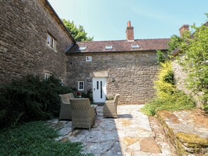 Un edificio de piedra con una puerta blanca, un pequeño jardín con muebles de mimbre y un camino de piedra.