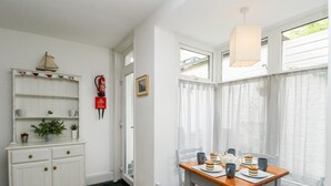 A dining area with a table set for four, a white cabinet with shelves, a fire extinguisher, and a window with sheer curtains. - Harbour Catch Cottage (Weymouth)