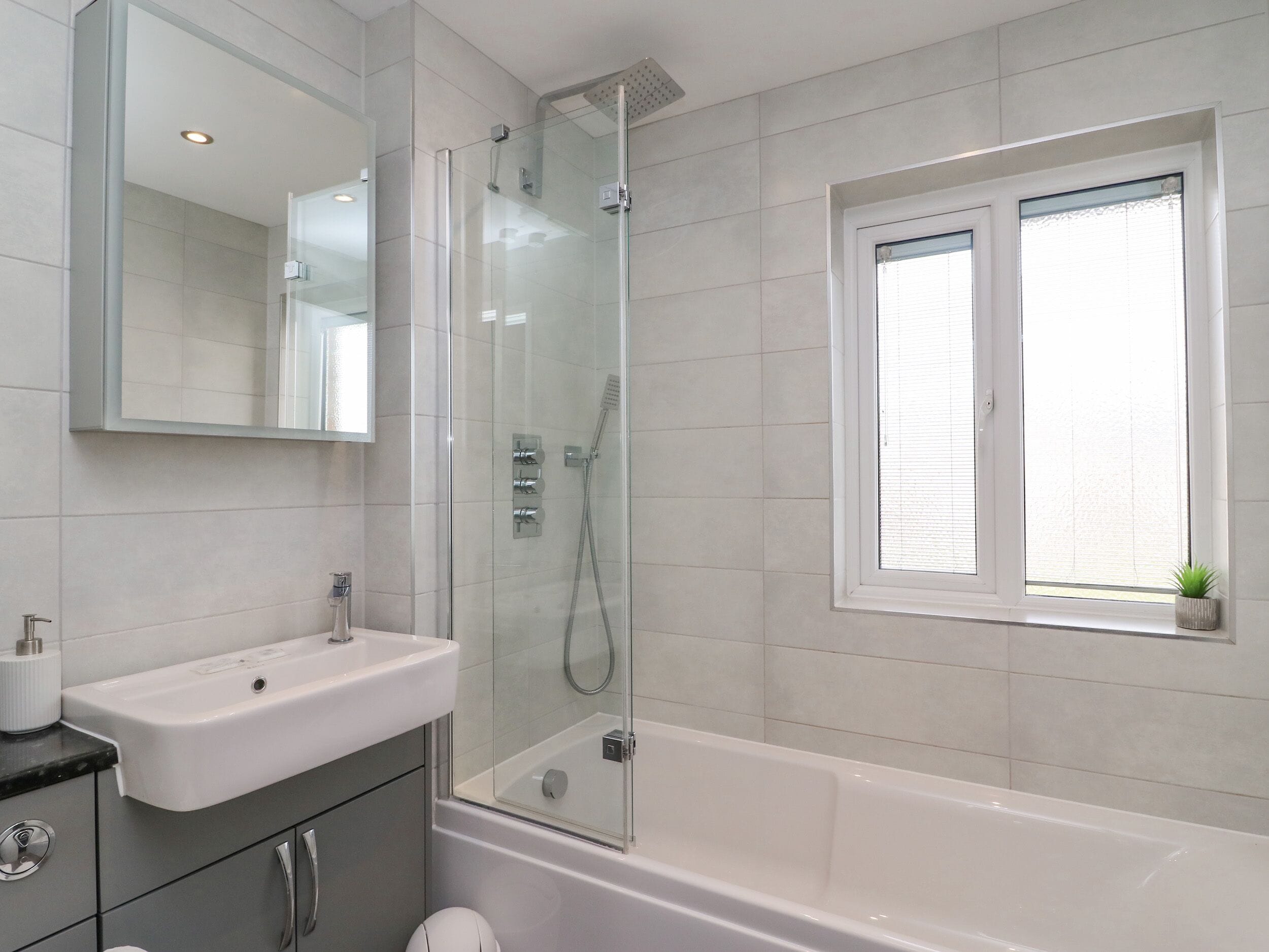 A modern bathroom with a glass-enclosed shower, a white bathtub, a sink with a mirror above, and a window with blinds.