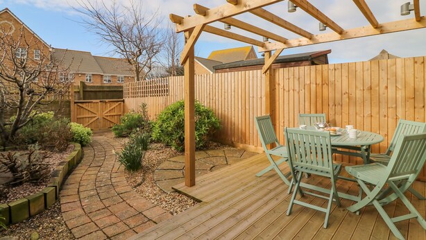 Una terraza de madera con una pérgola, una mesa y sillas, y un camino de ladrillos.