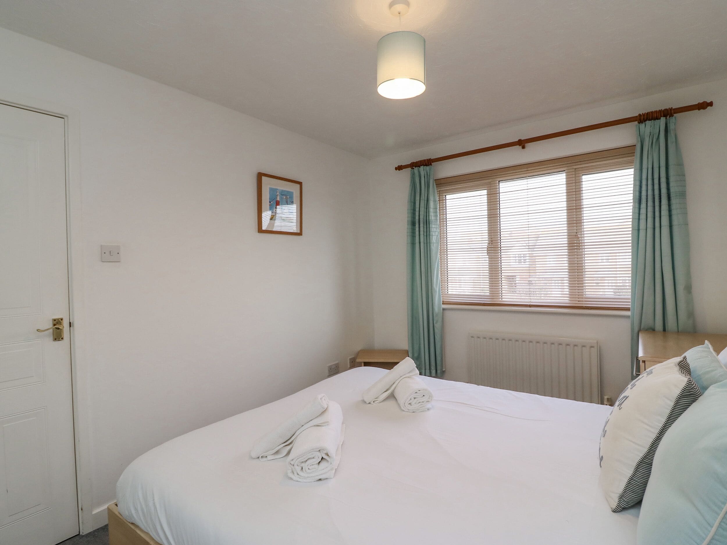 A hotel room with a bed, a bedside table, a lamp, a window with curtains, a radiator, and a door.