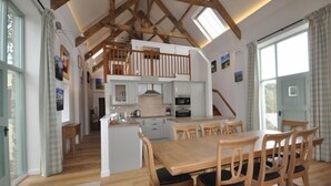 Un grand espace à manger avec une table et des chaises en bois, une cuisine avec des meubles blancs et une chambre à l’étage avec des poutres apparentes.