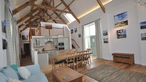 Un vaste espace de vie doté d’un plafond haut, de poutres en bois, d’une table à manger avec des chaises, d’un canapé et d’une cuisine à l’arrière-plan.