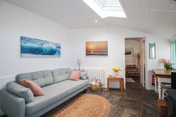 A living room with a grey sofa, a painting of a wave, a sunset painting, a lamp, a small table with flowers, and a view of the outside.