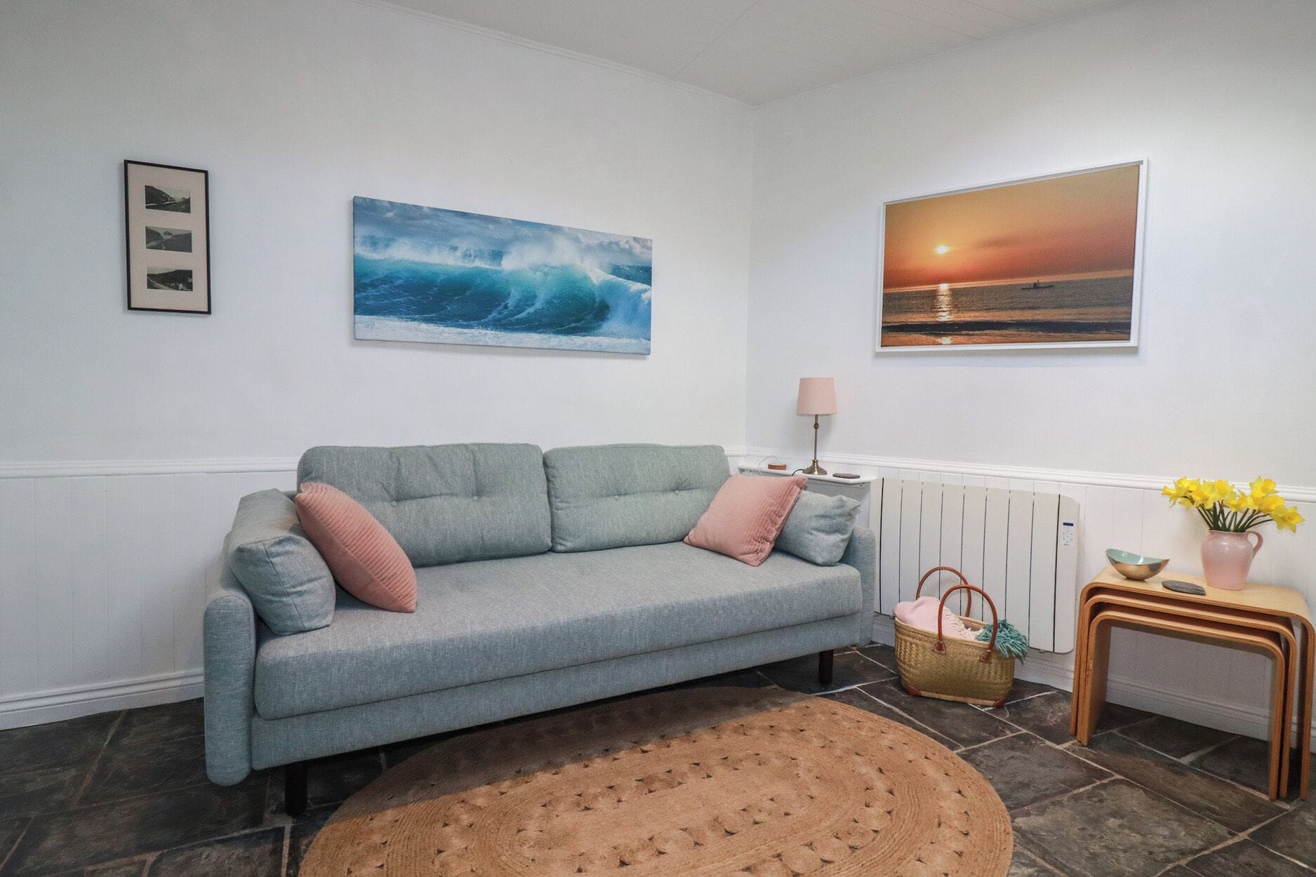 Una sala de estar con un sofá gris, un tapete redondo, una lámpara, un cuadro de un atardecer y un jarrón con flores.