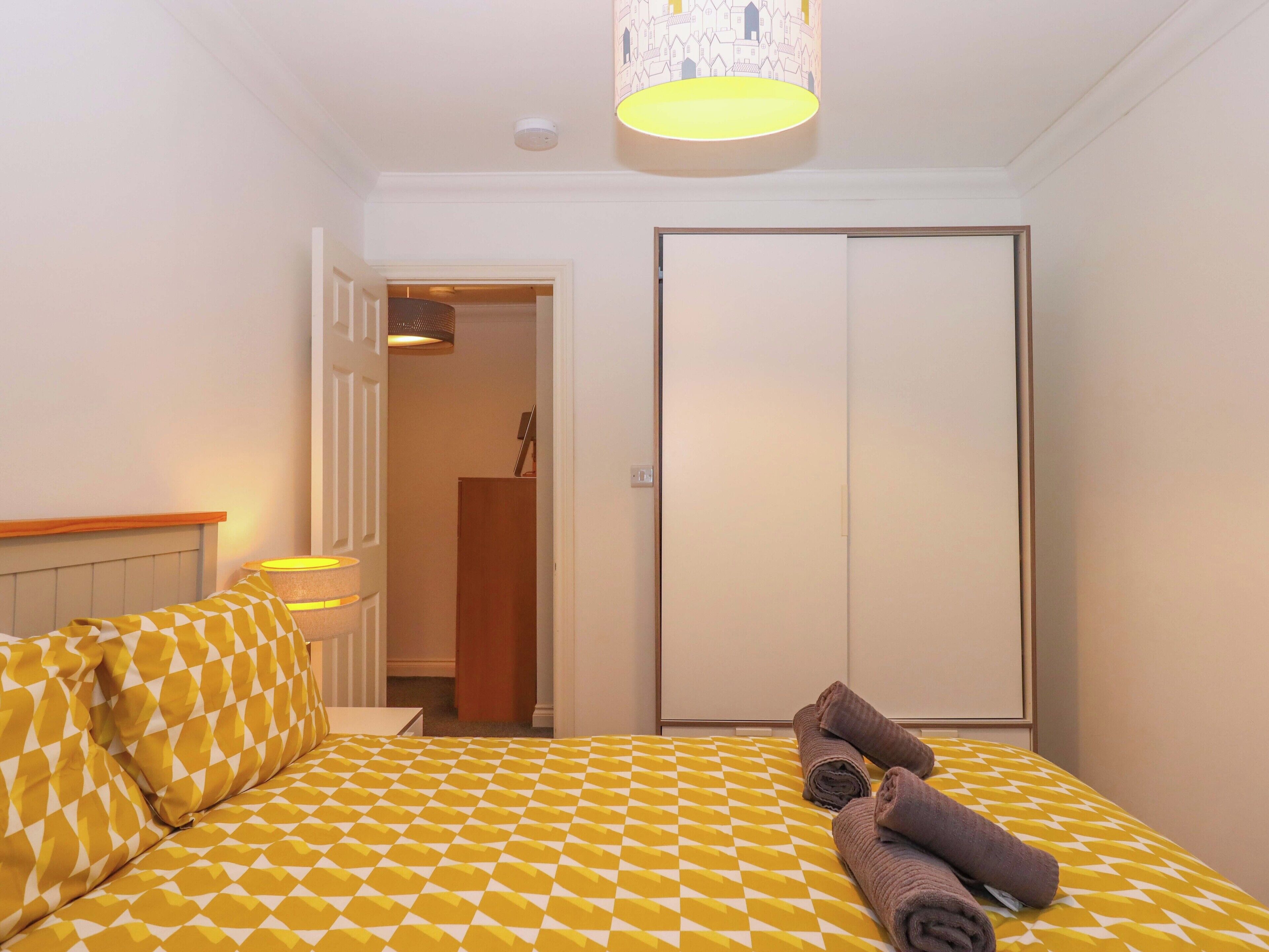 A hotel room with a bed featuring a yellow and white checkered bedspread, a wooden headboard, and a nightstand with a lamp. There is a door leading to a hallway and a closet.